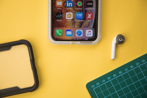 Top view composition of contemporary switched on mobile phone placed on yellow desk near phone cover and wireless earbud