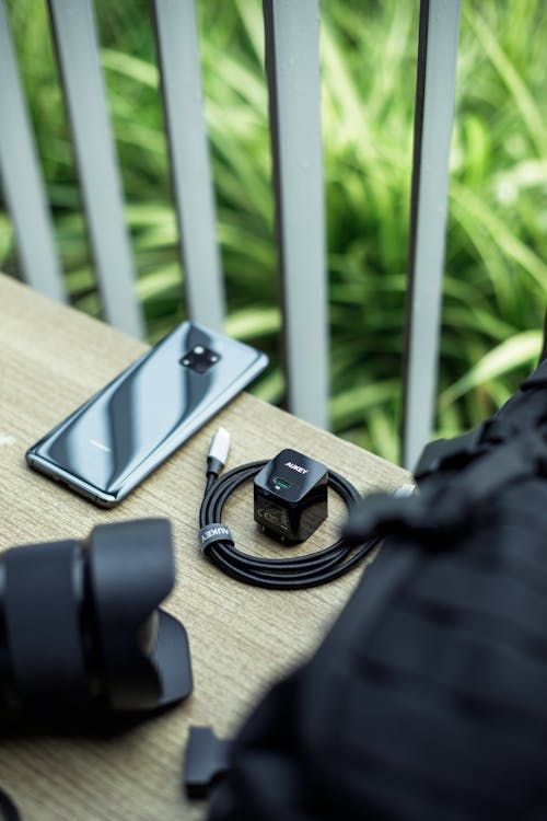 From above of contemporary mobile phone near USB connector and rucksack with photo camera lens on table near garden fence