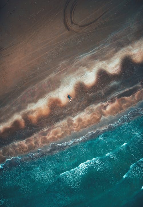 Breathtaking drone view of unrecognizable traveler lying on sandy beach of wavy turquoise ocean on sunny day