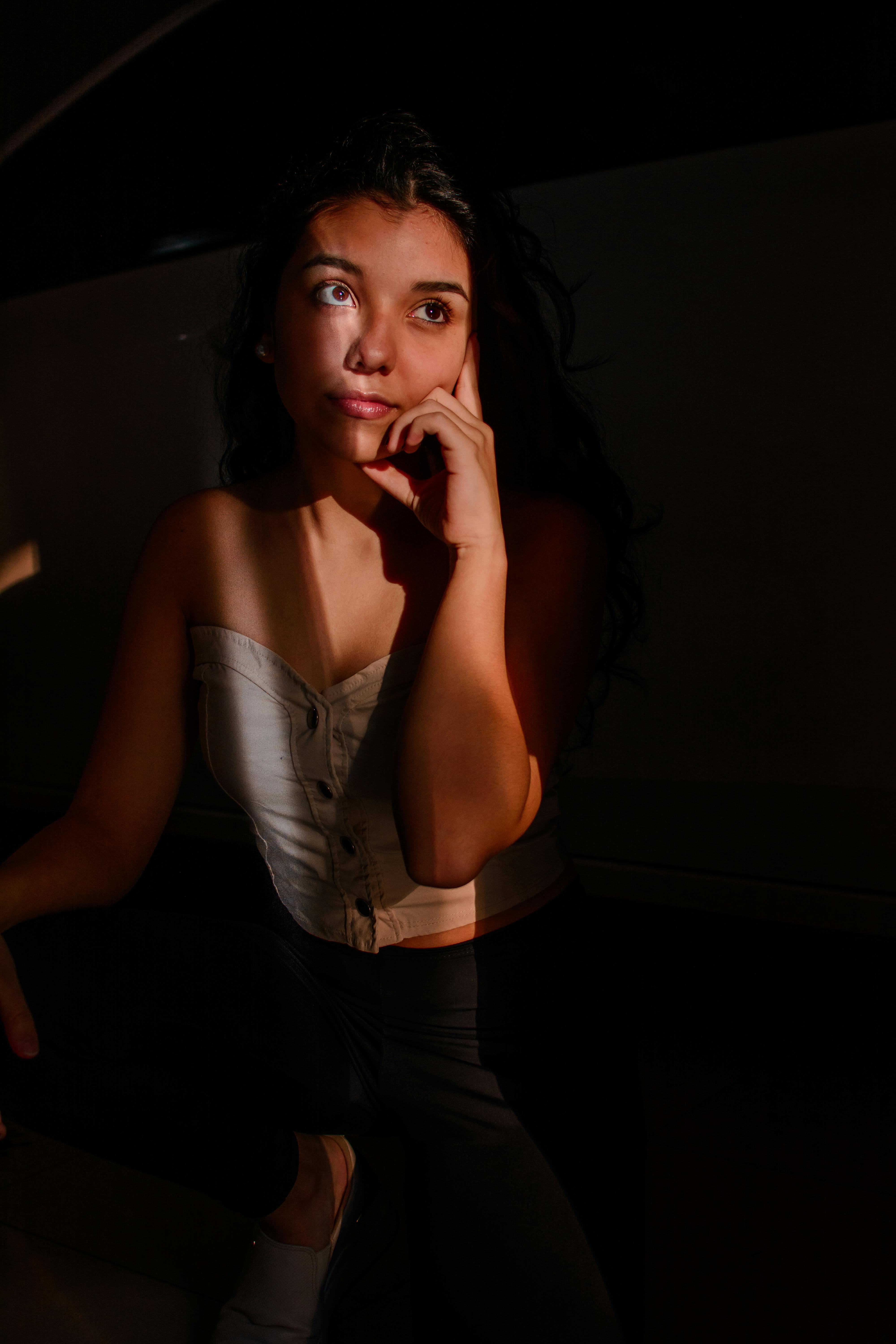 Pensive ethnic woman in trendy top leaning on hand while standing in sunbeam and looking calmly away