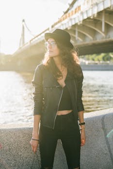 Young serious female in eyeglasses and fashion outfit standing near river and bridge while looking away