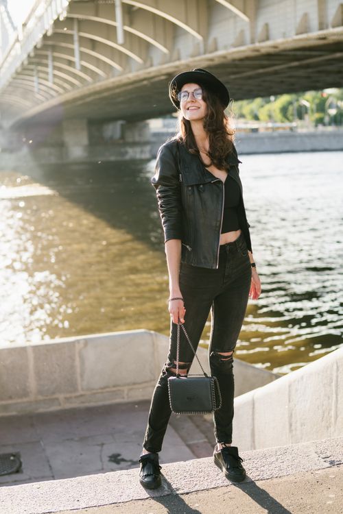 Free Full body smiling female in black trendy outfit and hat with handbag standing on top of street stair against city river under bridge in sunny day Stock Photo