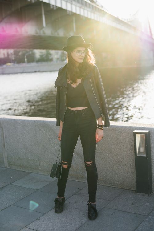 Free Full length trendy female in black outfit with jacket on shoulders standing alone on river embankment under city bridge in back lit Stock Photo