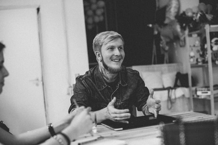 Cheerful Male Hipster Sitting At Table With Colleague In Workspace