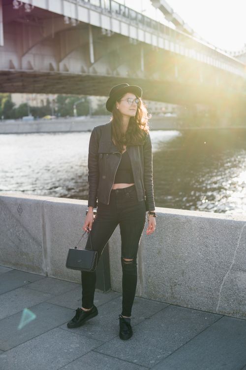 Free Full body stylish female in trendy black clothes with accessories standing alone on sidewalk of river embankment under city bridge in back lit Stock Photo