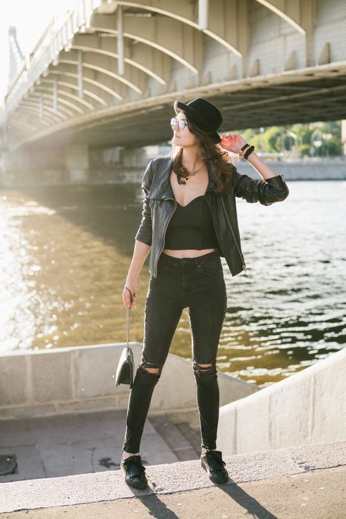 Free Full length of stylish young woman in black with accessories standing on top of stone stair leading to river flowing under bridge in back lit Stock Photo