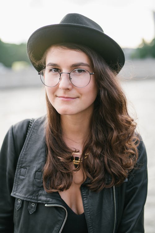 Stylish young female with loose hair wearing leather black jacket with hat and eyeglasses standing on street
