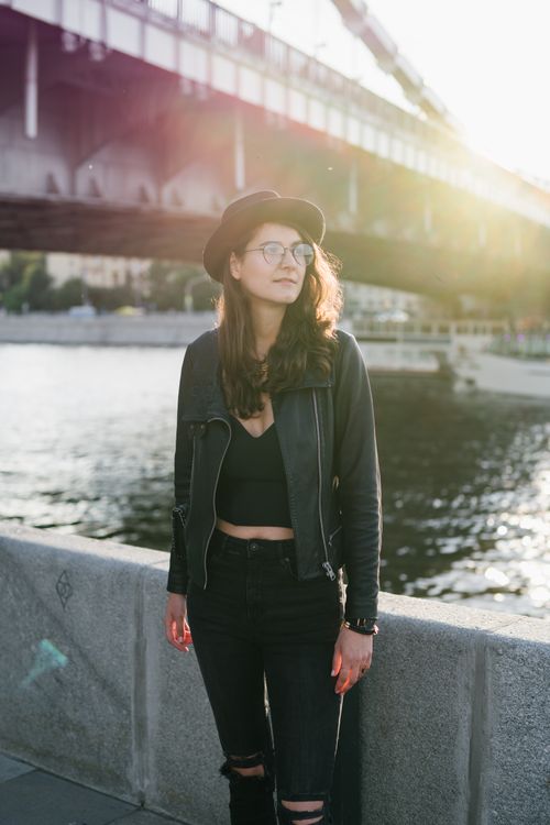Free Young female in black clothes with stylish accessories looking away admiring river while standing alone on embankment of river near city bridge Stock Photo