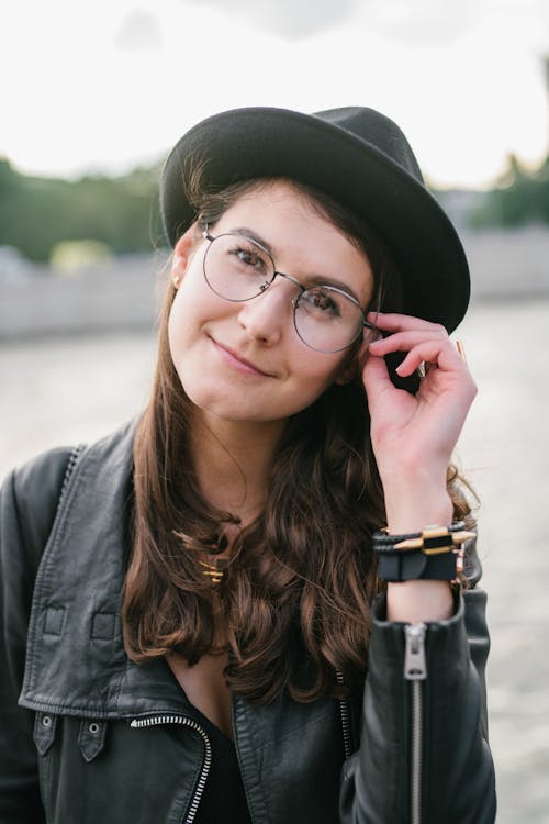 Optimistic young female in black jacket and hat adjusting glasses while standing on street of city against water