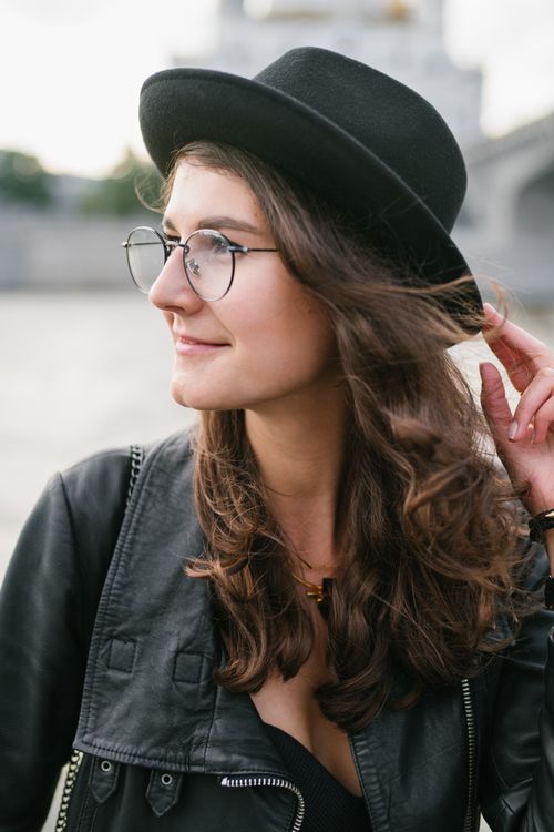 Free Young female in trendy clothes and hat and eyeglasses standing on city street in wind looking away positively Stock Photo