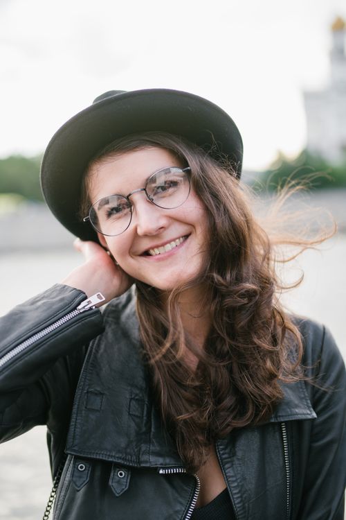 Free Positive young lady in elegant black jacket and glasses holding hat while standing in wind on city street Stock Photo