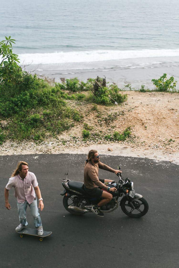 Friends Riding Motorbike And Skateboard Along Road Of Sea