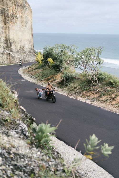 Free From above of unrecognizable male driver of motorcycle with skater riding behind on road and holding back park against mountain and sea Stock Photo