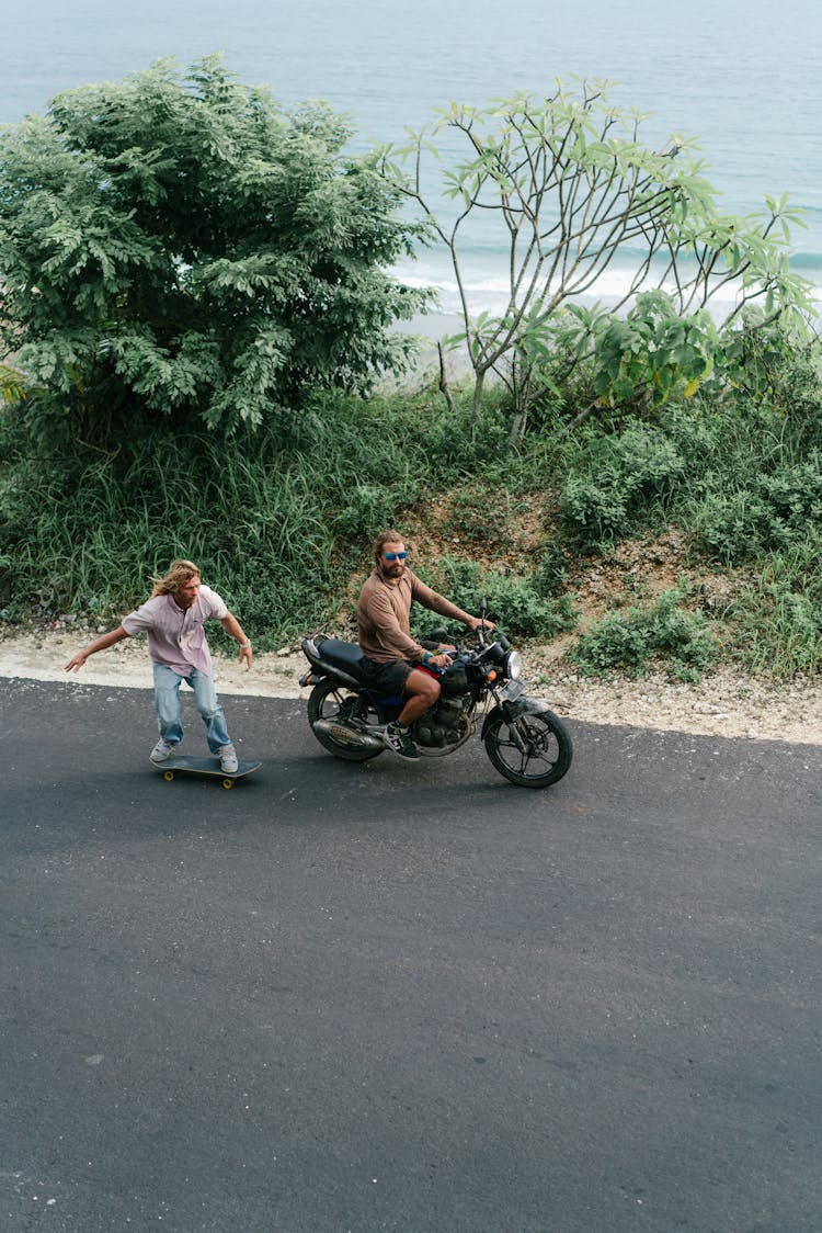 Man On Skateboard Riding After Motorbike On Road Of Shore