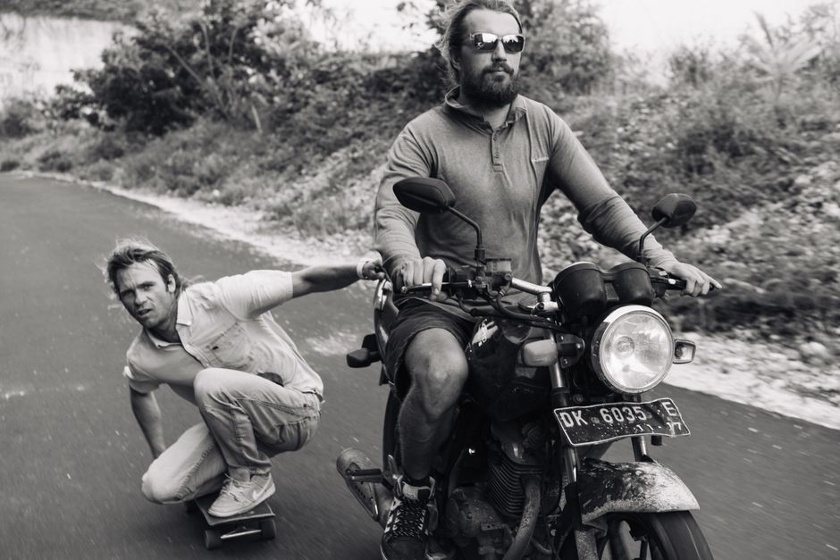 Black and white full length serious bearded biker riding motorbike with focused skater grabbing motorbike and being pulled on rural road