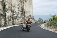 Full body man in casual wear riding skateboard while being pulled by biker on modern motorbike along asphalt road on picturesque seaside