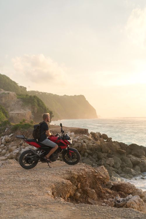 Free Side view full length faceless man sitting on modern bike parked on rocky seashore and enjoying amazing sea view during sunset Stock Photo