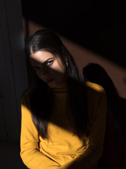 Free Portrait of serious young brunette in stylish yellow turtleneck sitting in dark room against brown wall and looking at camera provocatively while hiding half face in shadow Stock Photo