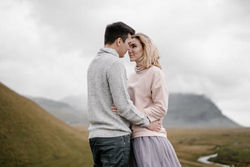 Free Side view glad young couple in warm sweaters embracing and looking at each other while spending time together on grassy hillside valley against majestic mountain and river on cold cloudy day Stock Photo