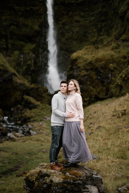Full length bonding couple in casual wear standing on rock in mountainous terrain against picturesque waterfall and looking away
