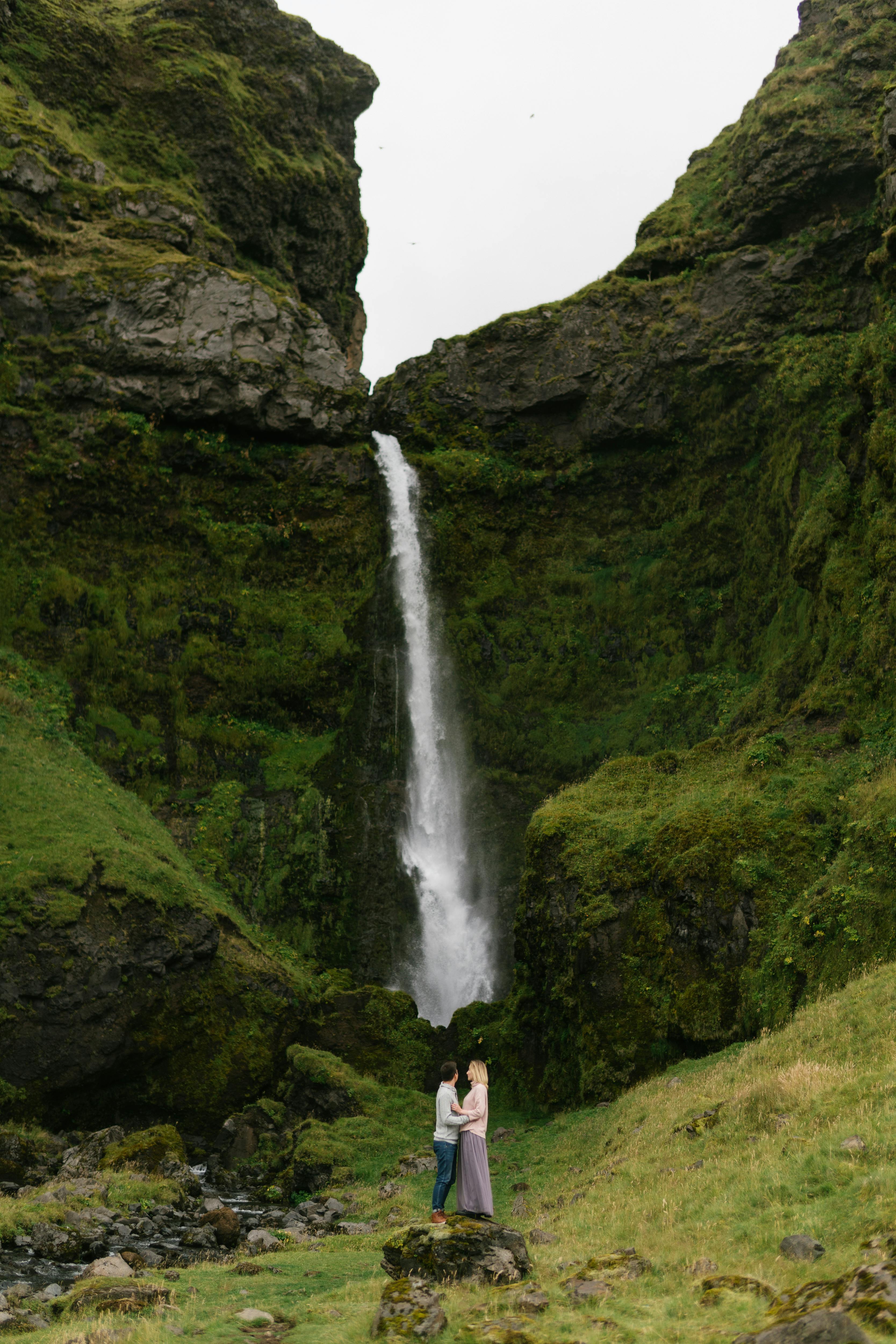 Side view anonymous couple standing on green grassy hillside and hugging while spending time together on mountain valley and admiring scenic waterfall