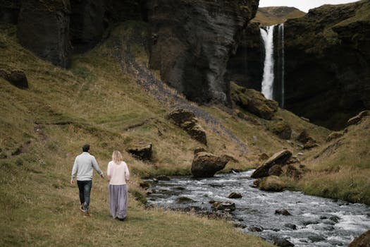 Back view full length faceless couple holding hands and walking along rapid mountain river towards waterfall while enjoying spectacular view of hillside