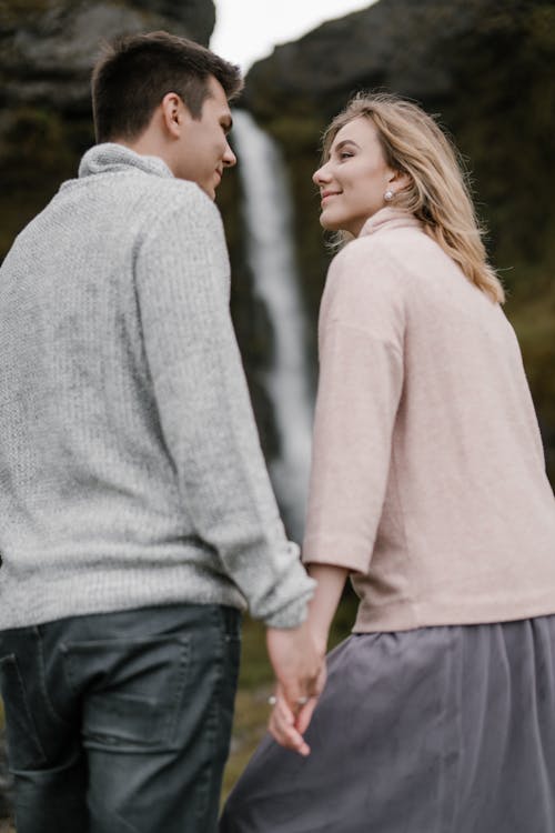 Low angle of loving young couple in casual outfits smiling and looking at each other against blurred cliff and waterfall