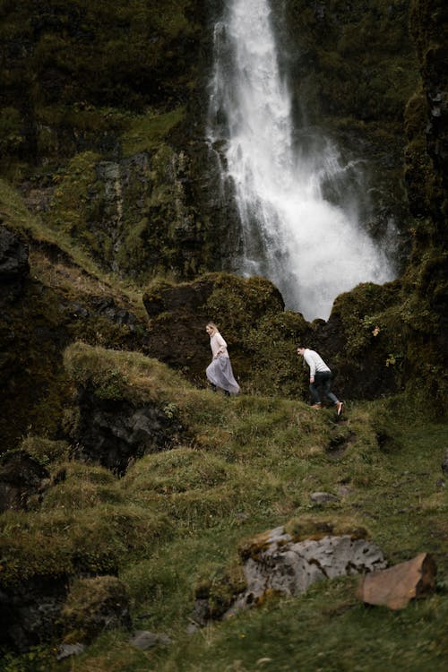 Side view of unrecognizable hikers climbing on rocky grassy cliff near powerful waterfall in Iceland