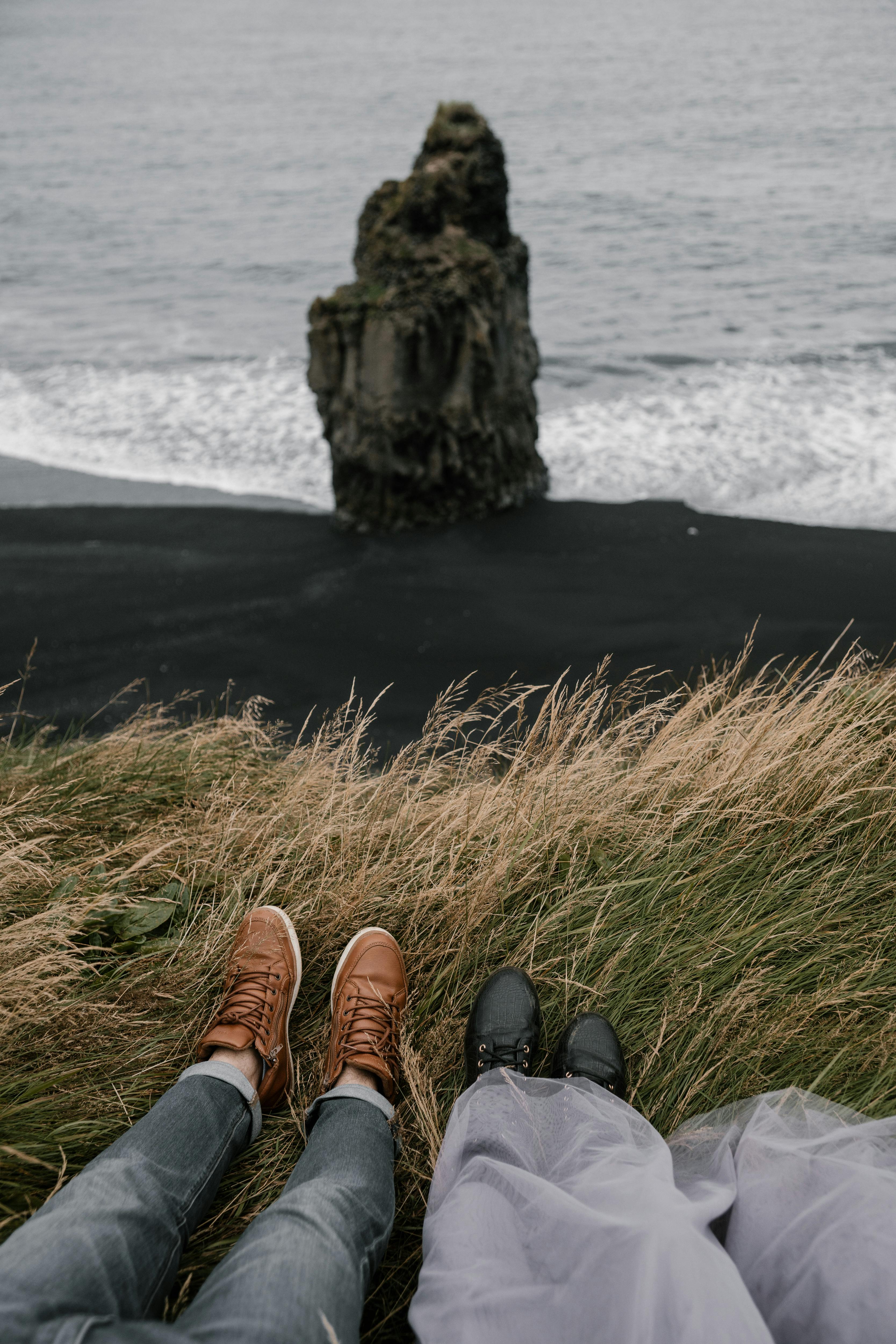 Crop anonymous boyfriend and girlfriend lying in withered autumn grass on black sand Reynisfjara beach and looking at small rock near sea waters edge on gray day