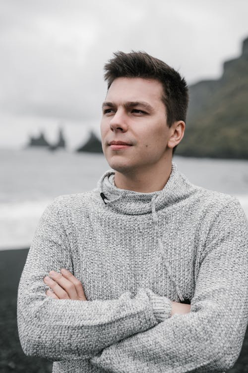 Young handsome short haired male in warm hoodie with arms crossed standing on sea beach and looking away on cloudy day