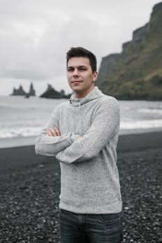 Young handsome man with short dark hair in warm gray hoodie standing with arms folded on cliffy seashore and looking at camera on overcast day