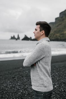 A young man in a sweater stands thoughtfully by a rocky seaside, embodying calm and introspection.