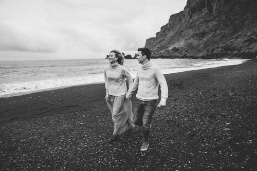 Black and white shot of romantic couple holding hands and running along gravel seashore while spending time together at seaside on chilly day