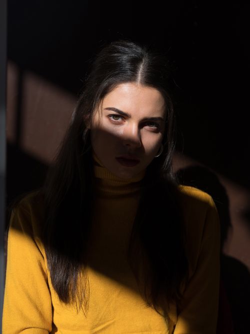 Free Portrait of serious young female wearing trendy yellow outfit with hiding half face in shadows and looking at camera alluringly while sitting in dark room against brown wall Stock Photo