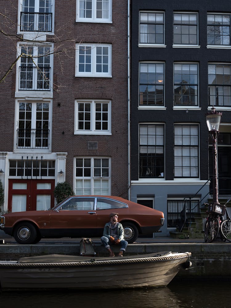 Man Sitting On City Riverside Near Retro Car And Boat