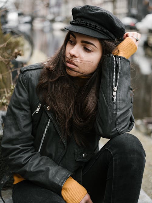 Stylish thoughtful young lady wearing leather jacket and trendy black cap while looking away pensively and leaning head on hand against blurred city background