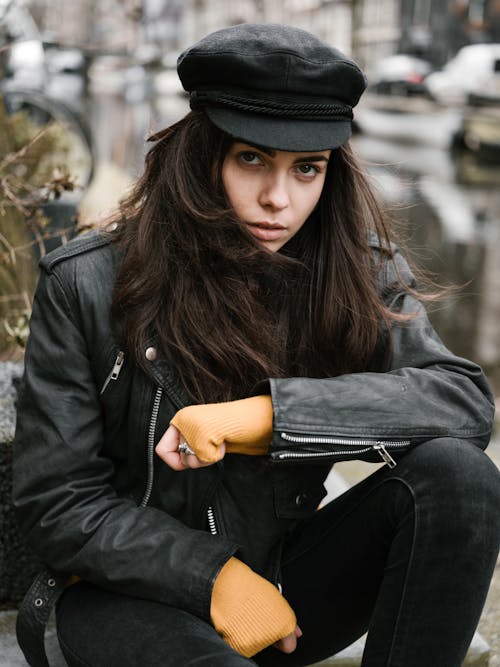 Fashionable young lady in leather jacket and trendy cap resting near old city canal on blurred background and looking at camera alluringly