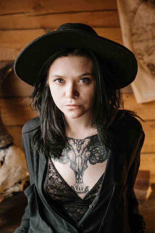 Serious young female in black lace dress and black shirt with trendy black hat on head and big tattoo on chest looking at camera in cozy vintage interior