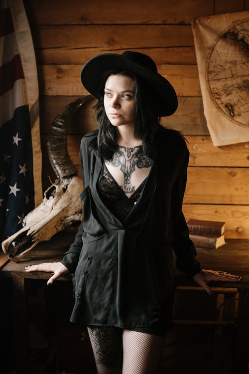 Confident young woman dressed all in black with big tattoo on chest looking away while standing near desk with steer skull and American flag in cozy vintage interior