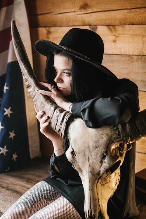 Stylish lady in black hat and short black skirt stroking long horn of steer skull and looking away while sitting on wooden desk with US flag in corner