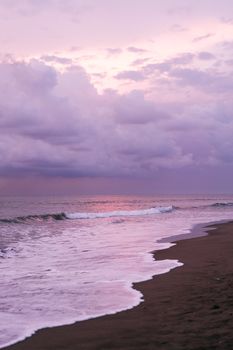 Amazing scenery of waving sea and sandy coast against picturesque cloudy purple sky at sunset time