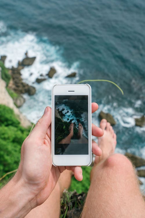 From above of anonymous barefoot male taking photo with mobile phone while sitting on high rocky cliff above ocean