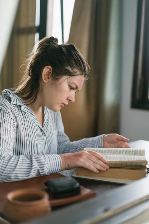 Concentrated young female in shirt searching information in textbook sitting at table at home
