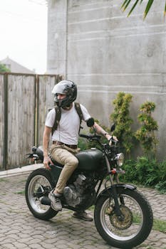 Male biker in casual clothes and helmet sitting on motorcycle and looking away in yard with closed gate in summer