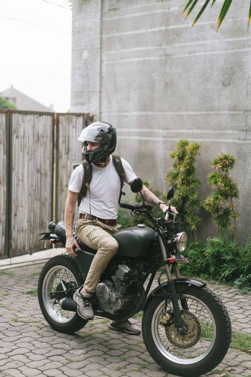 Free Male biker in casual clothes and helmet sitting on motorcycle and looking away in yard with closed gate in summer Stock Photo