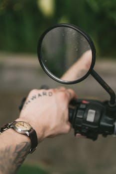 Crop anonymous male motorcyclist wearing wristwatch with tattoo saying ONWARD on hand grabbing handlebar partly being reflected in side mirror