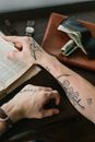 From above crop anonymous male with hands covered with tattoos looking through old reference books while sitting at brown table with leather wallet