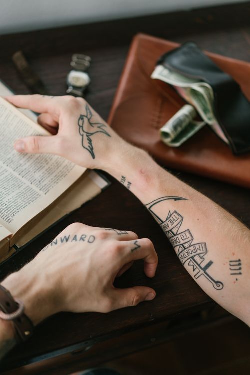 Free From above crop anonymous male with hands covered with tattoos looking through old reference books while sitting at brown table with leather wallet Stock Photo