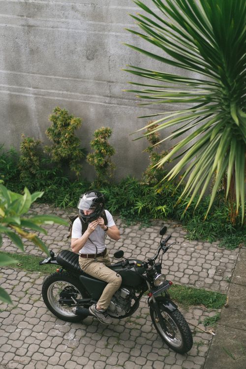 Free From above of male motorcyclist on black cafe racer in quiet yard with exotic greenery waiting for girlfriend to come and taking helmet off Stock Photo