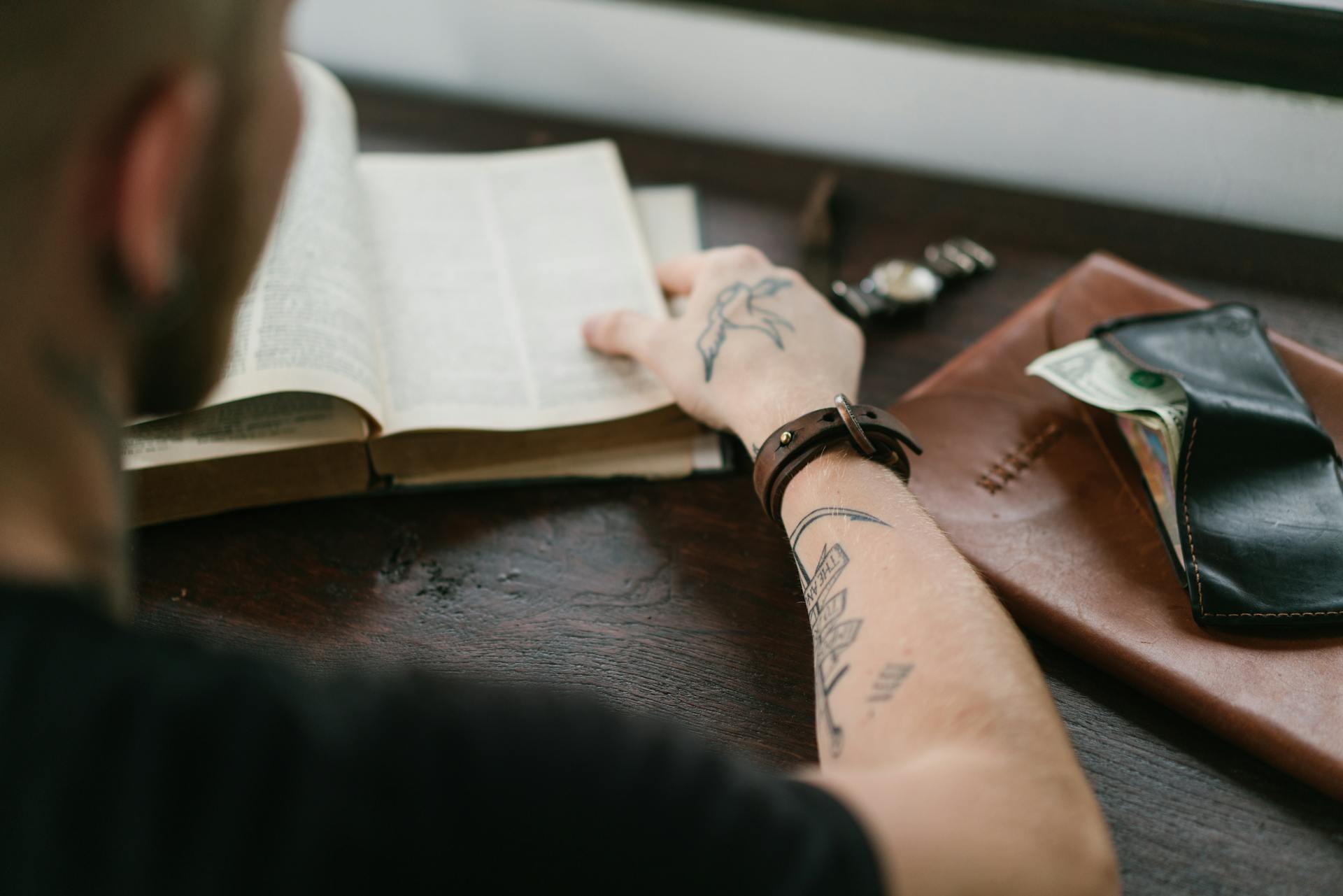 Back view of crop tattoed male student wearing elegant watch reading old books in library at desk with leather wallet and envelope